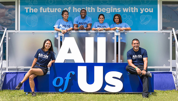 Six people sitting beside or standing behind 3D All of Us logo. Behind them is a banner that reads “The future of health begins with you.”