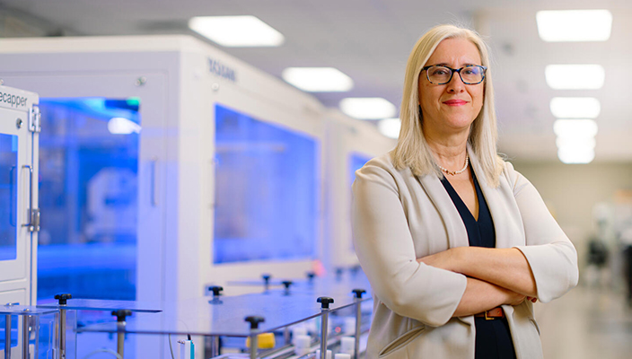 La doctora Mine Cicek está parada en el laboratorio y centro de procesamiento donde son almacenadas las muestras biológicas de All of Us. Está usando anteojos y sonriendo frente a la cámara.