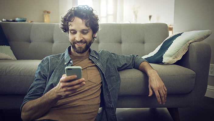 Un hombre adulto con barba está sentado en el suelo junto a un sofá, mientras lee algo en su celular, sonriendo. 