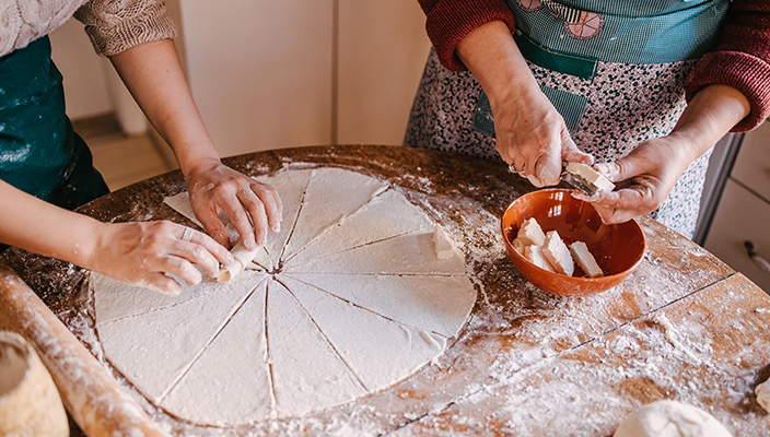 Imagen de las manos de dos personas paradas junto a una mesa cubierta de harina, preparando masa. Una persona está enrollando pedazos de masa previamente aplanada, y la otra está cortando otro ingrediente.