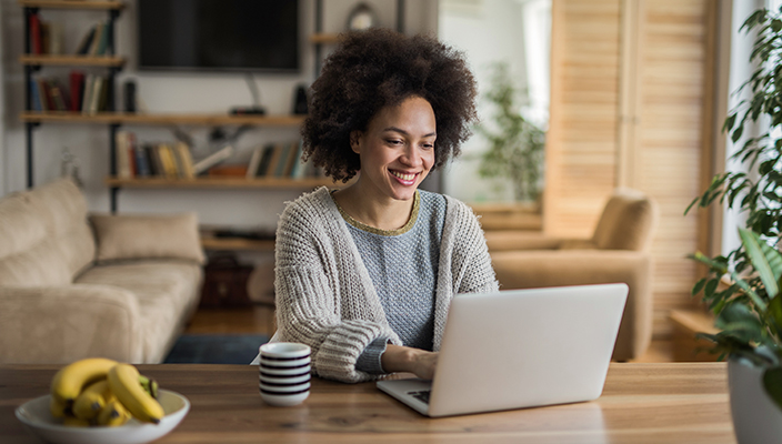 A woman is seated at a table in her house, using her laptop computer. She is smiling. There is a cup of tea next to her.