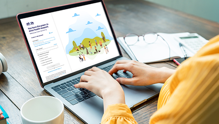 A person is working in a desk, typing in a laptop that displays a webpage related to All of Us. Reading glasses can be seen over documents next to the laptop.