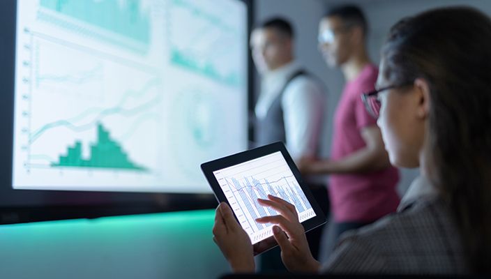 A woman wearing glasses is touching the screen of a tablet that displays a bar chart. In the background, a giant screen shows several graphics, next to 2 people who are analyzing them.